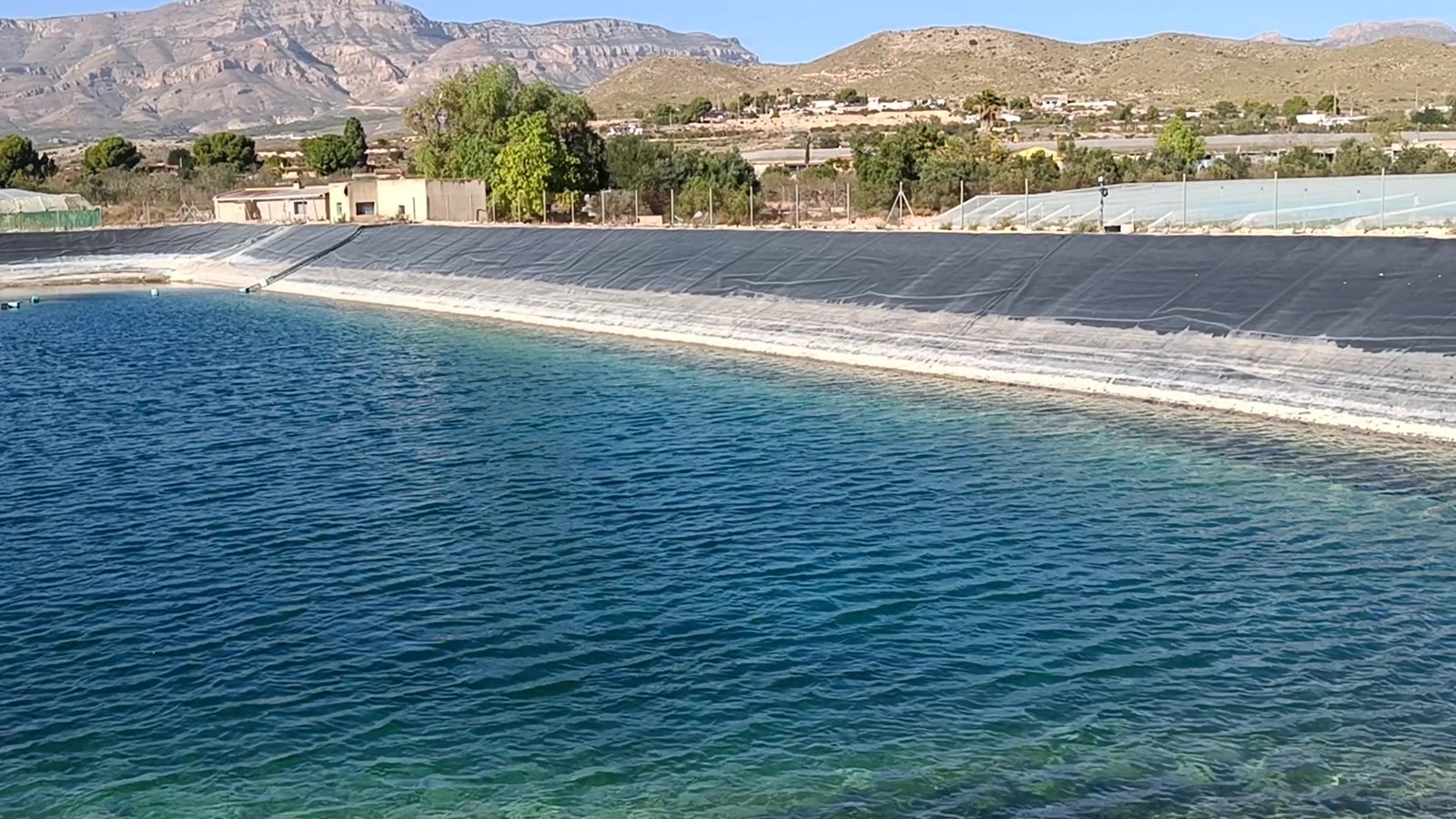Balsa de riego impermeabilizada con lámina, llena de agua, con invernaderos al fondo